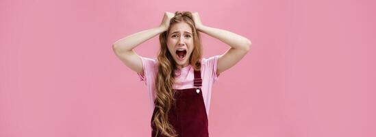Waist-up shot of woman in panic screaming and pulling hair out of head grimacing feeling anxiety from stress and problems standing bothered and distressed against pink background photo