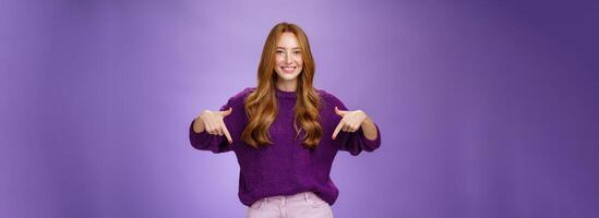 Girl giving recommendation to visit place, pointing down and smiling friendly with delighted and pleasant grin. Portrait of attractive redhead female model in purple warm sweater promoting copy space photo