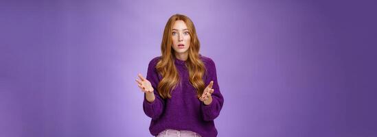 What wrong, I worried. Portrait of nervous and empathical young redhead woman feeling anxious raising hands questioned and looking at camera wondered expressing concern for friend over purple wall photo