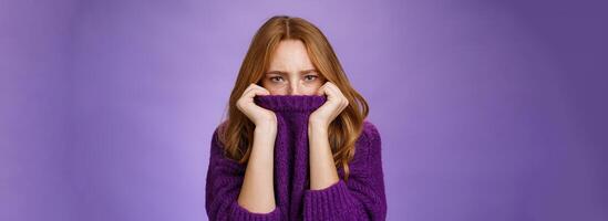 Gloomy upset redhead girlfriend pulling collar of purple sweater on nose, frowning and squinting offended, being aggrieved and resent, sulking moody and displeased over violet background photo