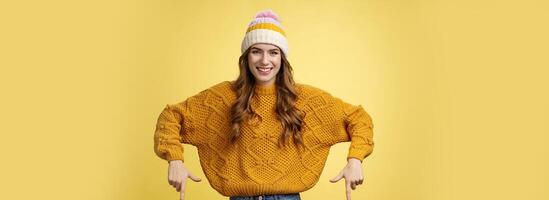 Friendly-looking helpful good-looking 20s college student girl wearing hat warm sweater smiling pleased showing you awesome promotion pointing index fingers down recommend bottom advertisement photo
