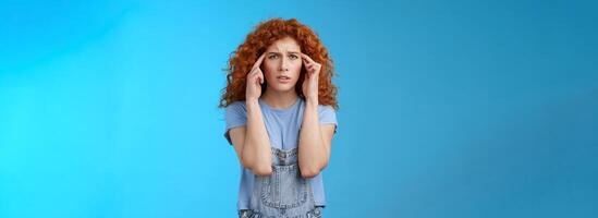 Confused panicking young distressed redhead curly girlfriend frowning touch temples cannot focus look perplexed worried not remember trying pull emotions together standing blue background photo