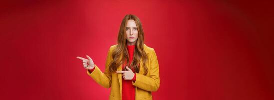Angry and offended disappointed redhead female in yellow coat frowning looking strict and serious at camera with dislike and agression pointing right asking explain yourself over red background photo