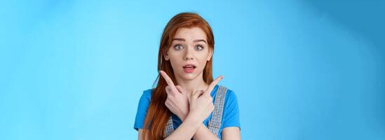 Questioned ambushed cute redhead woman asking advice picking product, cross hands pointing sideways, show left right choices, look camera wondered uncertain, making decision, blue background photo