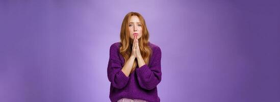 Sorrow and sad troubled redhead woman in warm sweater holding hands in supplication, pray gesture near chin frowning begging for mercy hoping for help or forgiveness with uneasy expression photo