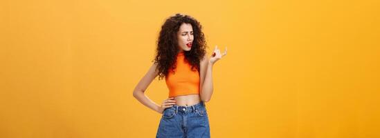 Portrait of moody arrogant and displeased european female with curly hairstyle and cropped top showing small and tiny object with fingers looking at little thing with disdain over orange background photo