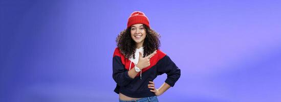 Studio shot of happy charming stylish city girl in beanie and warm sweatshirt ready rock winter up pointing at upper left corner and smiling delighted showing cool place over blue wall photo