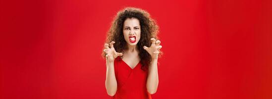 Woman like panter wanting scratch eyes of enemy. Portrait of pissed angry and pressured fed up woman clenching teeth and hands as if choking enemy with hate and anger in eyes being scary over red wall photo