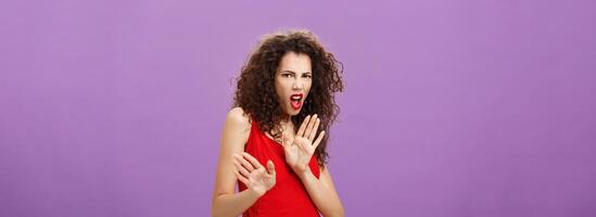 Disgusted arrogant good-looking female with curly hairstyle in red evening dress and makeup pulling palm in aversion and dissatisfaction grimacing rejecting suspicious offer over purple background photo