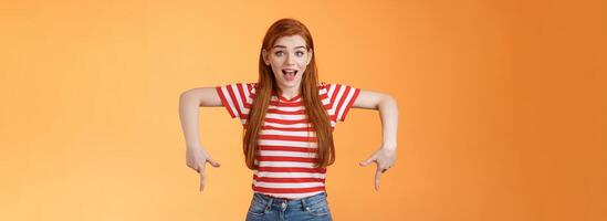 Enthusiastic lively cute redhead woman introduce incredible offer, pointing down, bottom copy space, smiling amazed, look camera, explain you awesome opportunity, feel excitement, orange background photo