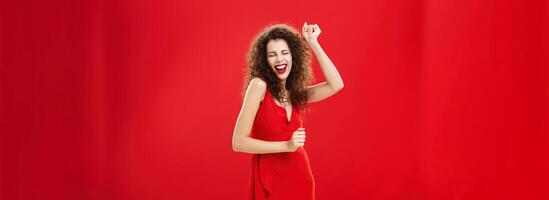 Rich woman forgetting all troubled on dance floor. Charming elegant young european female model in stylish evening dress moving to rhythm of music rejoicing raising hand, singing posing over red wall photo