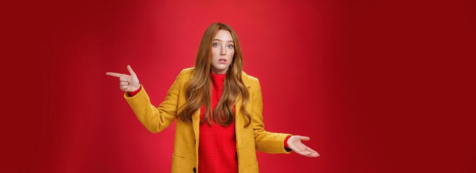 Woman missed client posing confused and unaware how happened shrugging making clueless gesture with palm as pointing right and looking questioned and uncertain at camera over red background photo