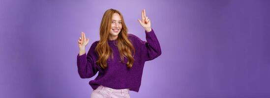 Girl in happy and energized mood dancing having fun making finger pistols gesture as smiling flirty at camera feeling thrilled and joyful at awesome party posing in purple sweater over violet wall photo