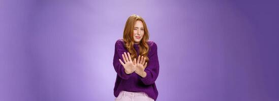 Gross take it away. Portrait of disgusted redhead woman step back and waving palms over body in refusal and no gesture grimacing and squinting from bad smell or aversion over purple wall photo