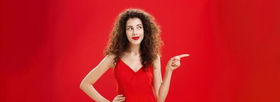 Stylish and elegant curious charming woman with curly hairstyle in red lipstick and trendy dress holding hand on hip pointing and looking left delighted and interested over studio background photo