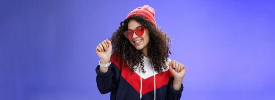 Stylish and feminine girlfriend dancing having fun feeling upbeat as liking weather feeling warm and cozy in stylish red beanie and sunglasses dancing with raised hands and broad smile over blue wall photo