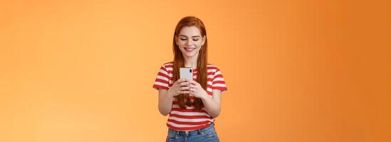 Friendly outgoing modern ginger girl communicating friends, chatting, send funny memes hold smartphone, smiling happy looking gadget display, use social media app enjoy new device feature photo