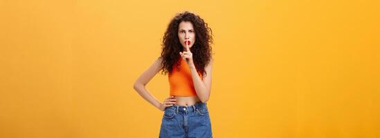 Sister displeased sibling saying cursing words standing serious and strict over orange background in stylish outfit shushing at camera saying shh with index finger over mouth demanding obey rules photo