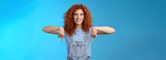 Charismatic sassy flirty redhead daring ginger girl curly haircut pointing down index fingers smiling broadly enthusiastic explore new store pointing promo like cool advertisement blue background photo
