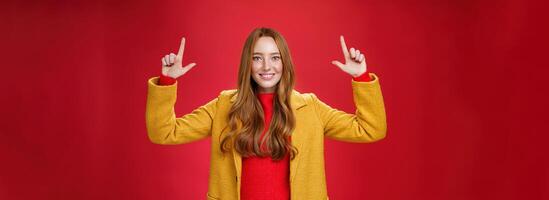 Indoor shot of redhead attractive woman in yellow fall coat raising hands promoting advertisement as pointing up and smiling broadly with satisfied pleasant expression over red background photo