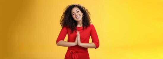 Welcome come inside. Portrait of friendly and polite good-looking female host in red dress with curly hair holding hands in namaste gesture tilting head and smiling as inviting guests in asian style photo
