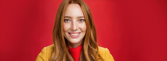 Headshot of hopeful and happy young redhead 20s girlfriend with cute freckles and blue eyes smiling broadly with faithful excited expression having high hopes, posing over red background photo