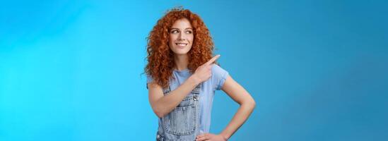 Cheerful motivated charismatic good-looking happy smiling redhead 25s curly-haired woman summer dungarees pointing upper left corner look aside pleased show promo suggest try-out photo