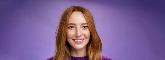 Headshot of charming attractive and happy young redhead woman with freckles and bright white smile, grinning satisfied at camera as posing over purple background friendly and delighted photo