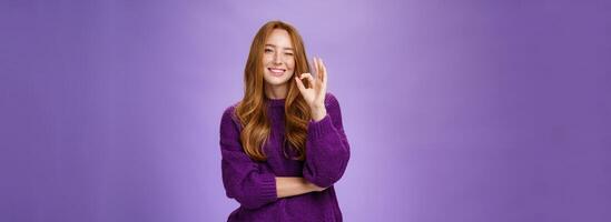 Nice work you did great. Satisfied and happy charming assertive and supportive redhead woman in purple sweater smiling and winking in approval showing okay gesture, liking product over violet wall photo