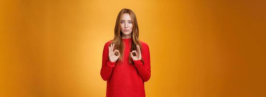 Calm and confident girl showing self-assurance and professionalism as making okay gesture with unemotional expression assuring everything goes by plan standing over orange background, giving approval photo