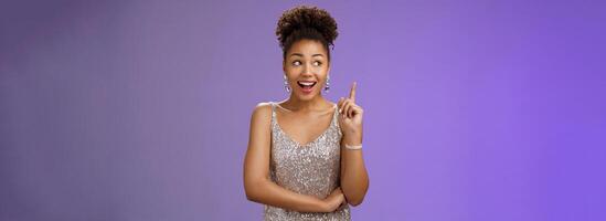 Creative joyful dreamy young african-american girl in silver dress raise index finger eureka gesture look aside inspired have awesome idea sharing suggestion tell plan aloud, standing blue background photo