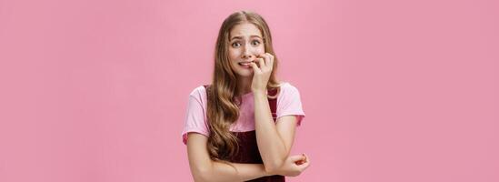 Woman panicking and overreacting feeling nervous biting fingernails and staring afraid at camera trembling from fear over pink background posing in cute dungarees with natural wavy hairstyle photo