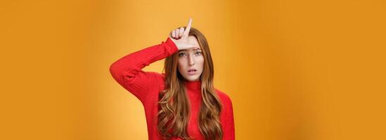 Arrogant and self-satisfied redhead woman humiliating person showing loser sign on forehead mocking and disdain rival standing confident and snobbish over orange background photo