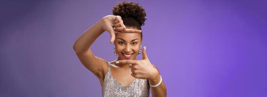 Creative good-looking african woman in silver glittering dress searching inspiration make finger frame look camera pleased smiling satisfied found perfect photo angle, standing blue background