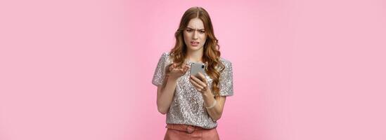 Frustrated pissed woman keep receiving strange messages cringing grimacing bothered look smartphone display gesturing irritated confused, cannot understand article, pink background photo