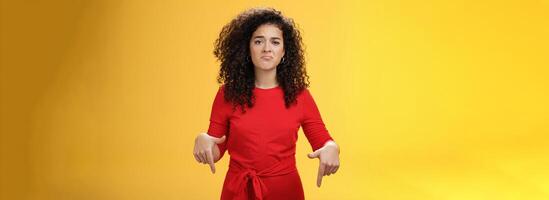 Portrait of gloomy girl with sad face pursing lips down frowning disappointed and pointing down with regret and unsatisfied expression, missing opportunity feeling upset over yellow background photo