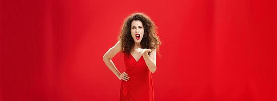 How dare you betray me. Pissed outraged bossy and displeased european adult woman with curly hairstyle in elegant dress bending towards camera with raised palm, complaining blaming and judging photo