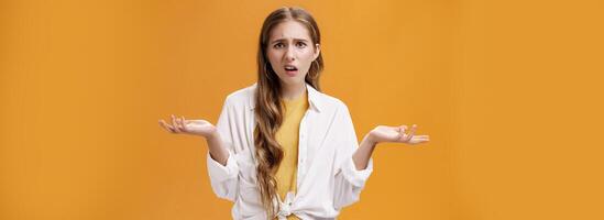 What, I do not understand. Portrait of arrogant questioned impolite girl being pissed with questiones shrugging with hands spread sideways making irritated grimace standing confused over orange wall photo