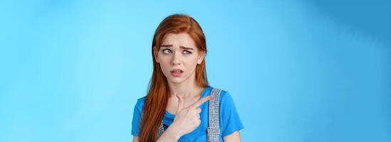 Stupified redhead hesitant girl show aversion, pointing looking left reluctant, full disbelief, stare with dismay uncertain about product, disliking strange weird guy standing blue background photo