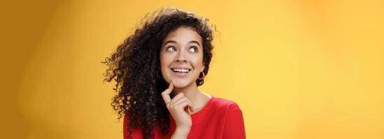 Dreamy girl making assumptions what gets as christmas present looking curious and happy at upper left corner, thinking, touching chin, standing thoughtful with desire and wish in mind over yellow wall photo