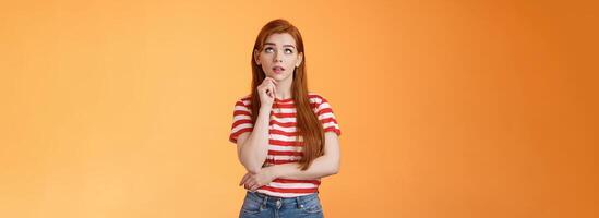 Thoughtful cute curious redhead woman looking intrigued up, touch chin thinking, making assumption, observe interesting copy space product, stand orange background pondering information photo