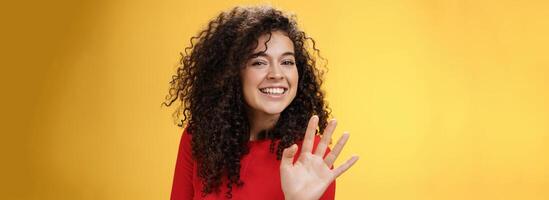 Charming friendly and self-assured attractive curly woman waving cute with palm to say hi or hello smiling broadly greeting man trying flirt in party posing joyful over yellow background photo