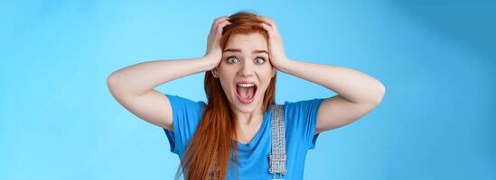 Shocked redhead girl shouting panic, feel pressured deadlines, scared see mess, grab head, scream frightened distressed, stare camera hopeless, anxiously react losing lottery, blue background photo