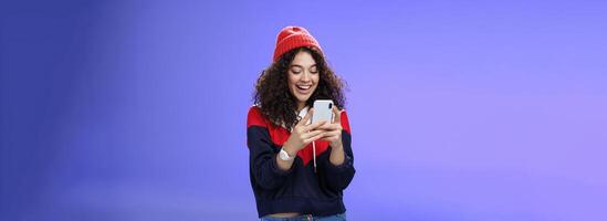 Joyful and sociable stylish caucasian woman with curls in warm beanie having fun laughing and chuckling as using smartphone typing funny message looking at mobile phone screen happily over blue wall photo