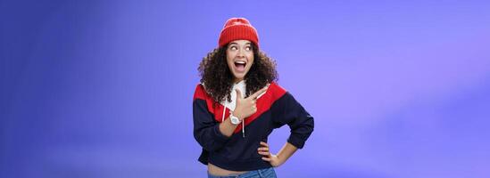 Cute and playful girl in winter stylish outfit and watch smiling broadly with excitement and joy pointing, looking at upper left corner thrilled and joyful as posing over blue background photo