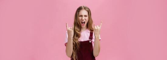 Cute and excited party girl with wavy hairstyle in corduroy overalls yelling yeah and showing rock-n-roll gesture having fun enjoying hard music being daring and rebellious over pink wall photo