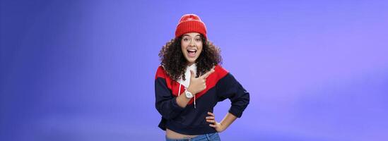 Portrait of enthusiastic and sociable european woman in warm hat and sweatshirt smiling delighted with amused grin as pointing at upper left corner impressed and astonished with awesome promotion photo