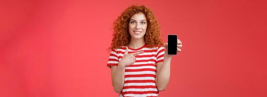 How about this app. Redhead curly friendly cute girl help friend find oufit online store hold smartphone pointing phone screen smiling broadly recommend social media photo filter, red background