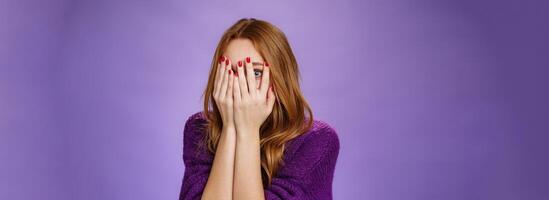 Waist-up shot of curious and cute redhead female covering face with palms from fright as watching horror movie peeking through fingers with interest of what happen next over purple background photo
