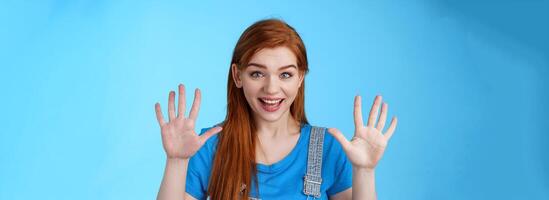Close-up friendly joyful redhead caucasian girl, long ginger hairstyle, raise hands show number ten, counting dozer, explain fingers, raise arms surrender, smiling joyful, stand blue background photo